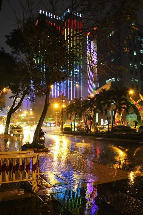 Ảnh thành phố về đêm mưa với tòa nhà đèn màu phản chiếu trên đường ướt